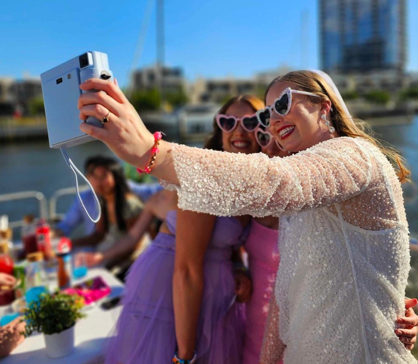 The bride in her veil and heart sunglasses taking a selfie with her girls on deck, the city behind them