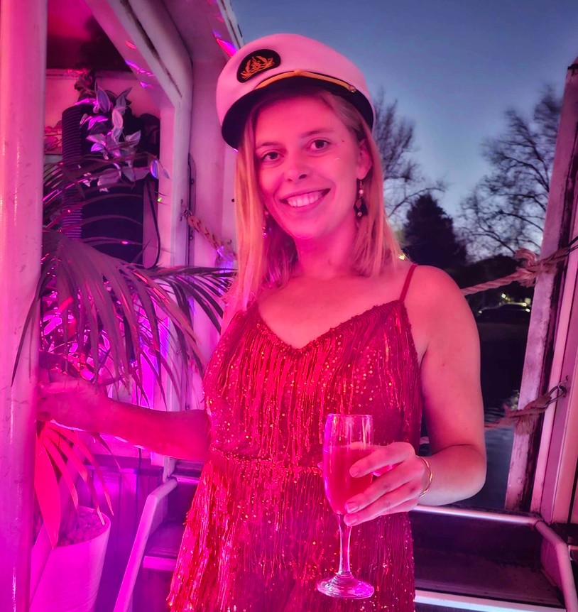 A hen in a captain hat and red sequins with a glass of bubbles, the boat lit pink at dusk