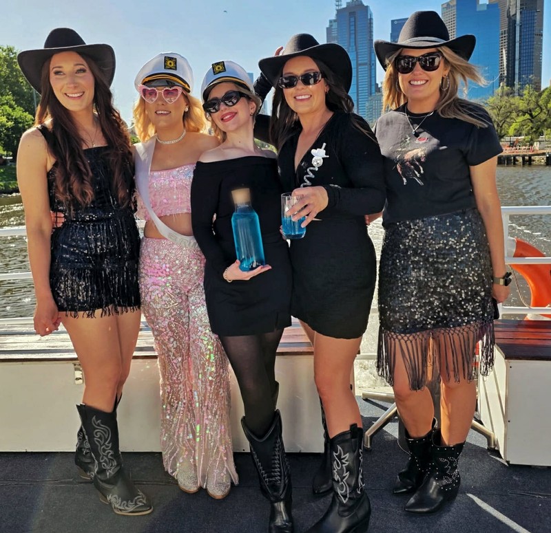 A hens crew mixing cowgirl hats and captain hats on the deck of a river cruise, the city towers behind them
