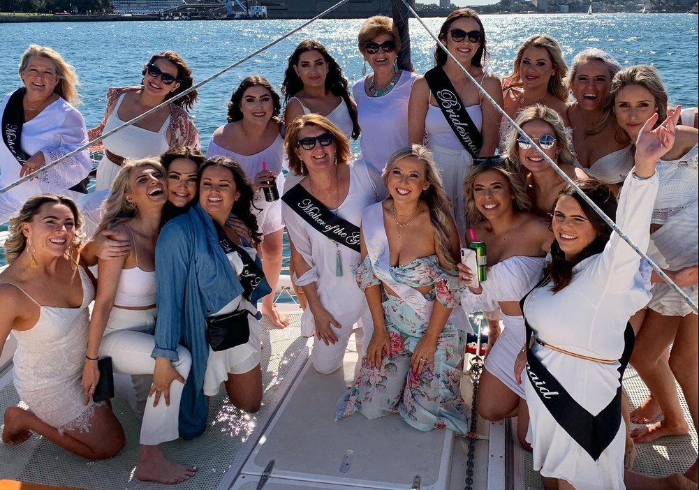 A hens crew in white on a catamaran, the mums in their sashes kneeling beside the bride, the harbour sparkling behind