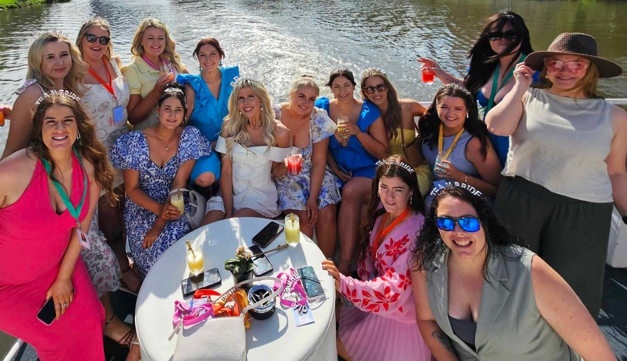 A big hens crew around a round table on deck in the sunshine, drinks in hand and the river behind them