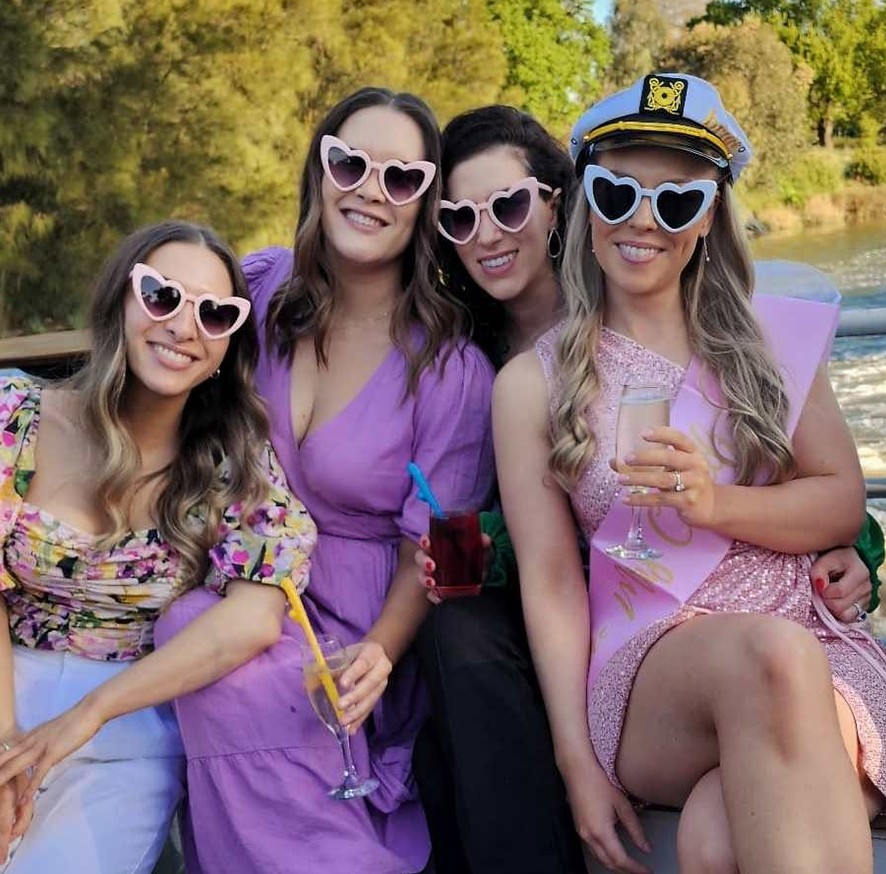 The bride in a captain hat and heart sunglasses with three friends on a river cruise, glass in hand