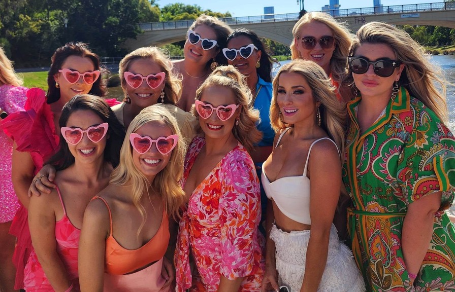 A hens crew in pinks, oranges and heart sunglasses on deck, a city bridge behind them