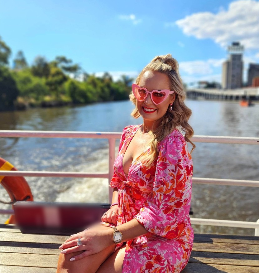 A hen in pink heart sunglasses and a floral dress sitting on the back deck, the river and the city behind her
