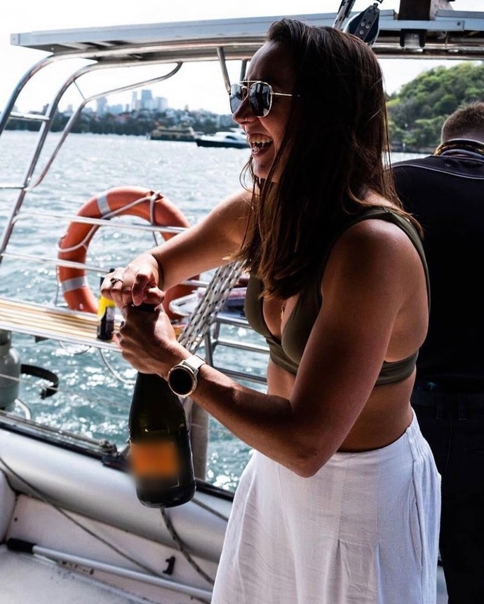 A hen laughing as she pops a bottle of bubbles on the deck, the harbour behind her