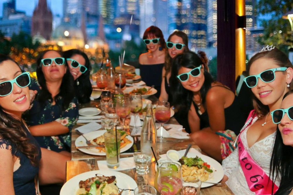 A hens group in sunglasses at a long dinner table with the city behind