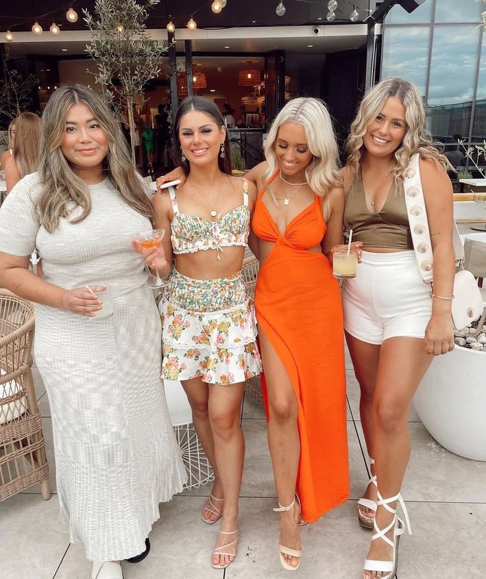 Four friends in florals, orange and white summer dresses with drinks on a rooftop