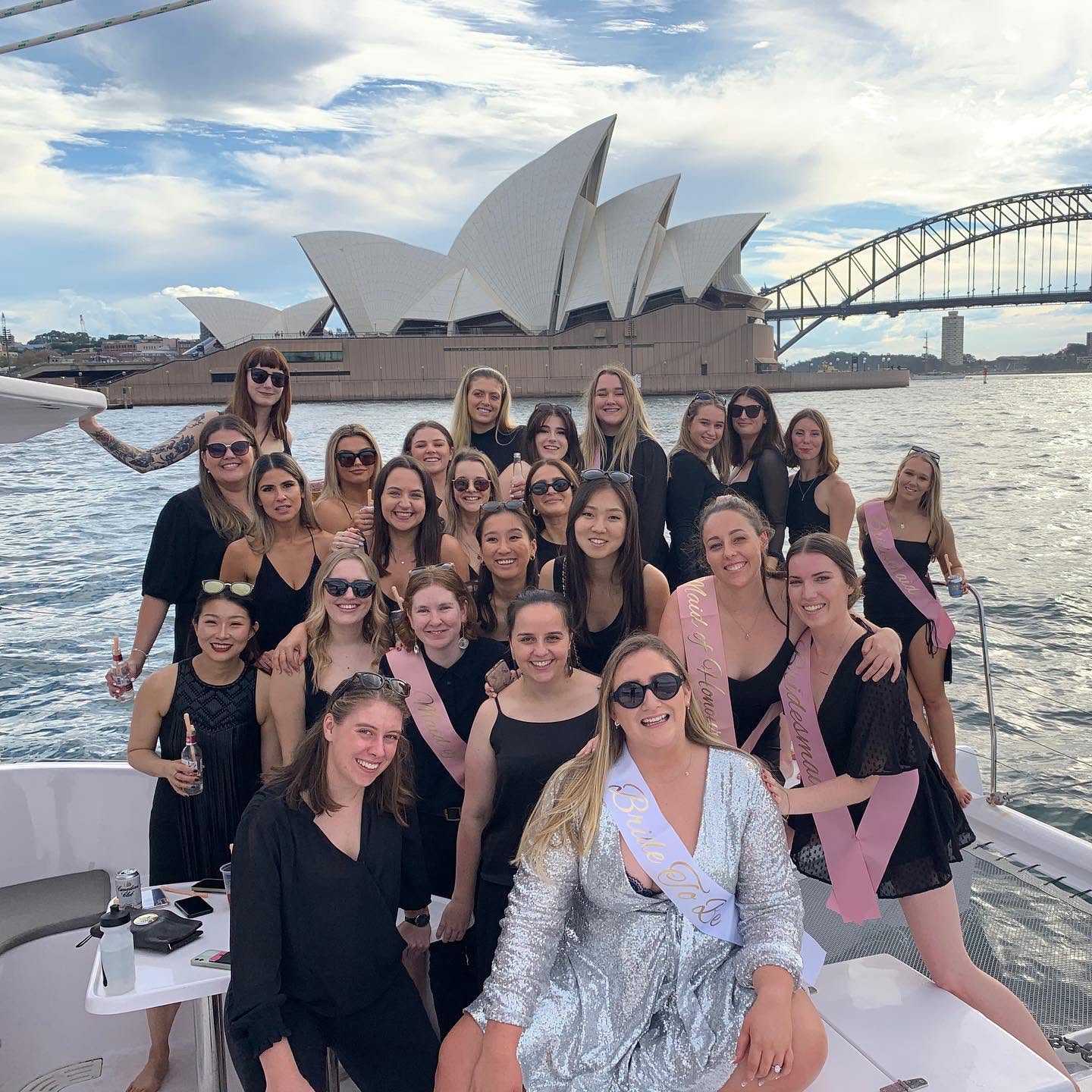 A big hens crew in black on the deck of a catamaran on the harbour, the bride in her sash in the middle, the Opera House and the bridge behind them
