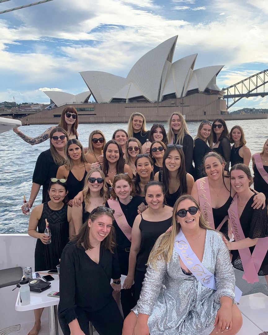 A big hens group in black, the bride-to-be in her sash, on a catamaran with the Sydney Opera House and Harbour Bridge behind