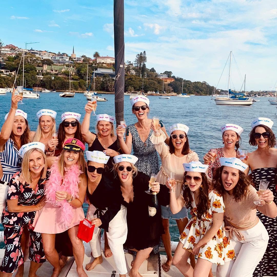 A hens crew in sailor hats toasting with bubbles on the deck of a yacht, moored boats behind