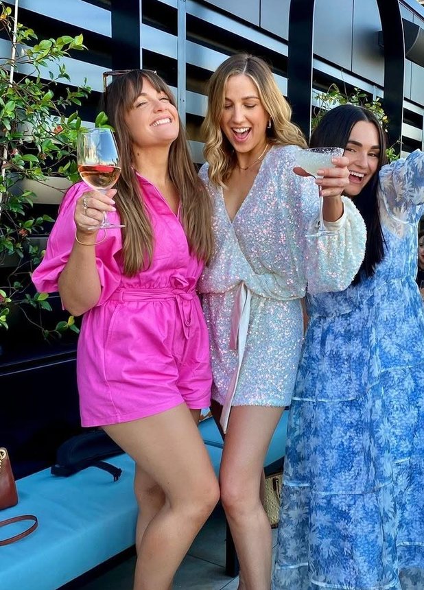 Three friends in pink, white sequins and blue florals raising their glasses on a rooftop