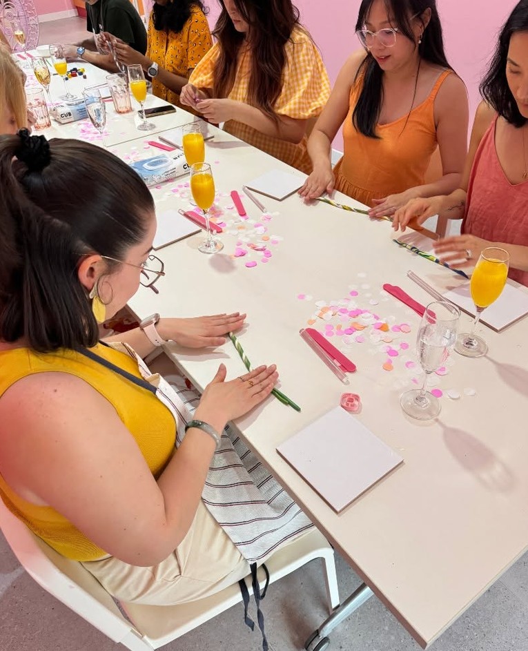Friends at a long table with glasses of orange juice and bubbly and pink confetti, rolling striped clay on white tiles
