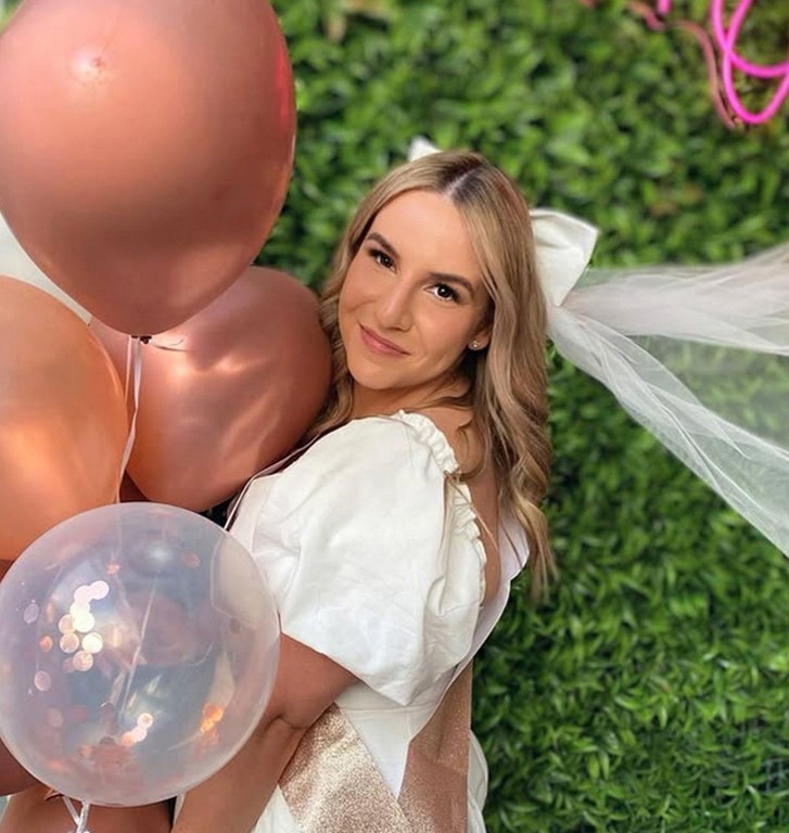 The bride in a white dress and bow veil with rose gold balloons in front of a green wall