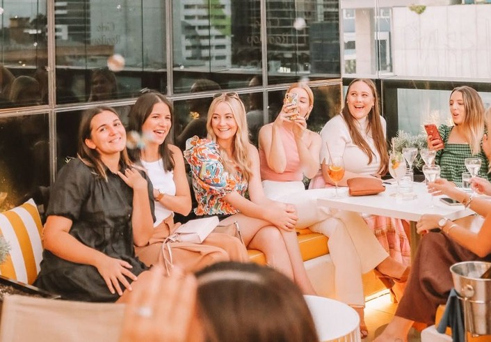 A hens crew laughing together on a rooftop lounge with drinks as bubbles float past