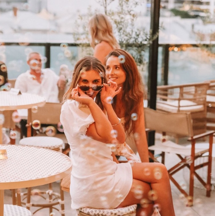A friend in a white dress peering over her sunglasses as bubbles float around her on a rooftop at sunset