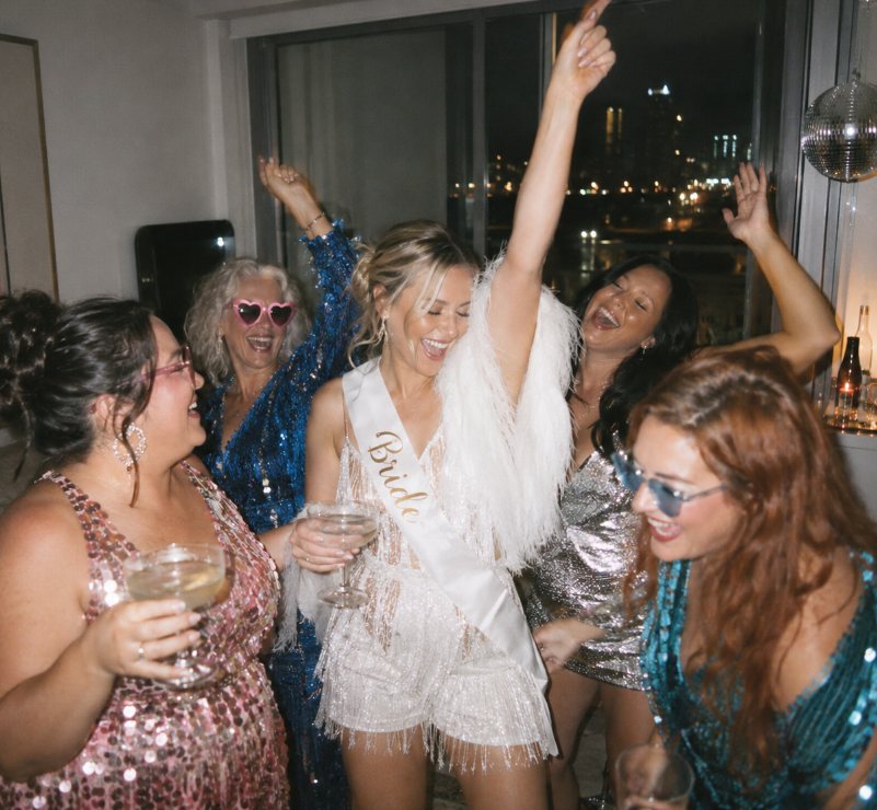 A bride in a white fringed outfit and feathers dancing with her arm up, friends of every age in pink, blue and silver sequins dancing around her, a disco ball and city lights behind