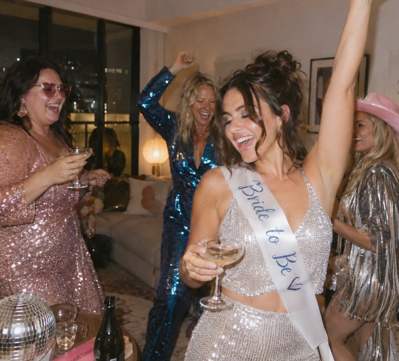 A bride to be in a silver sequin two-piece and sash dancing with her arm up, friends in sequins and a pink cowboy hat behind her