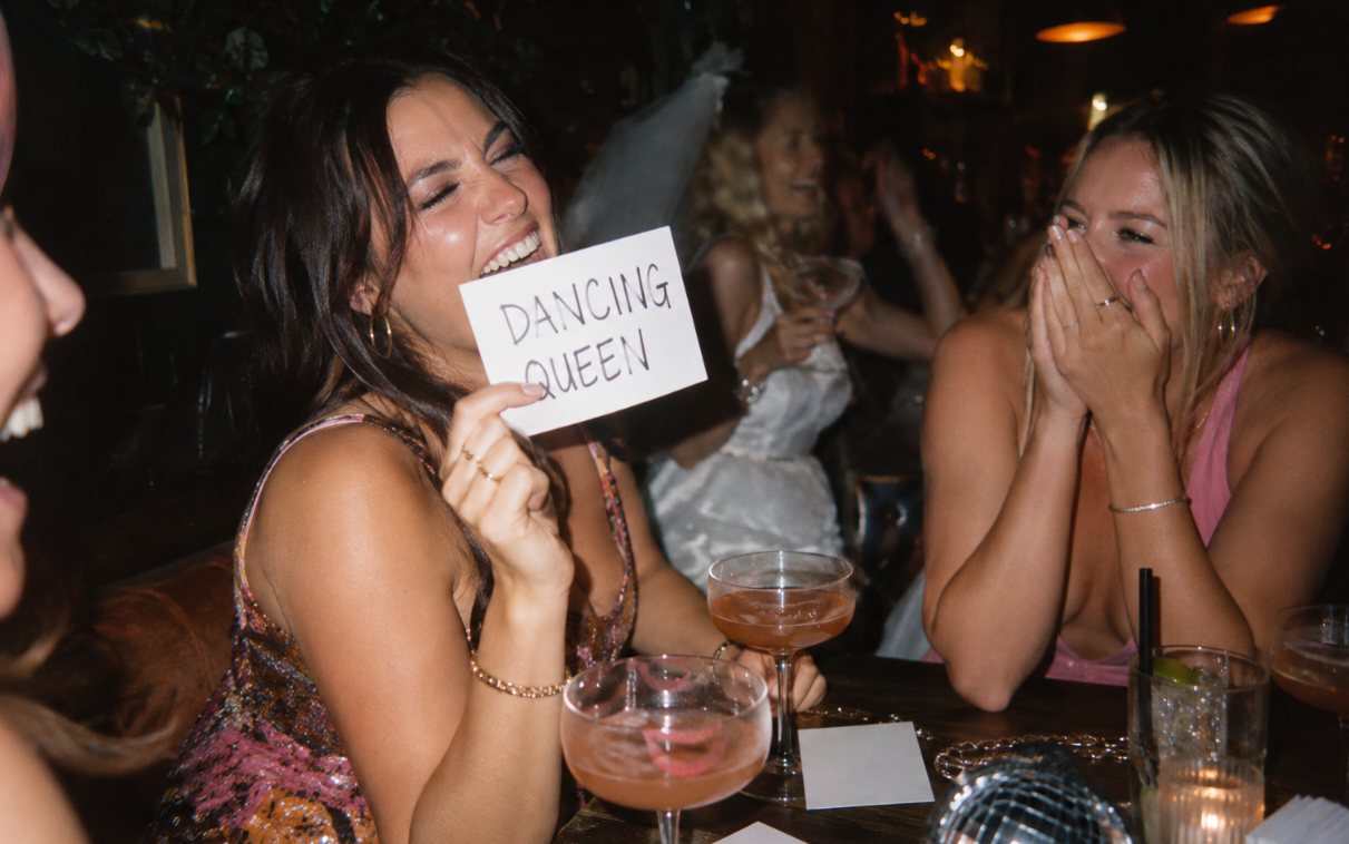 Friends laughing around a bar table, one holding up a handwritten Dancing Queen card, another hiding her face behind her hands, the bride in white behind them