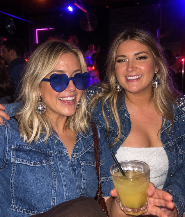Two friends in denim jackets and disco-ball earrings, one in blue heart sunglasses, smiling at a bar with a drink in hand