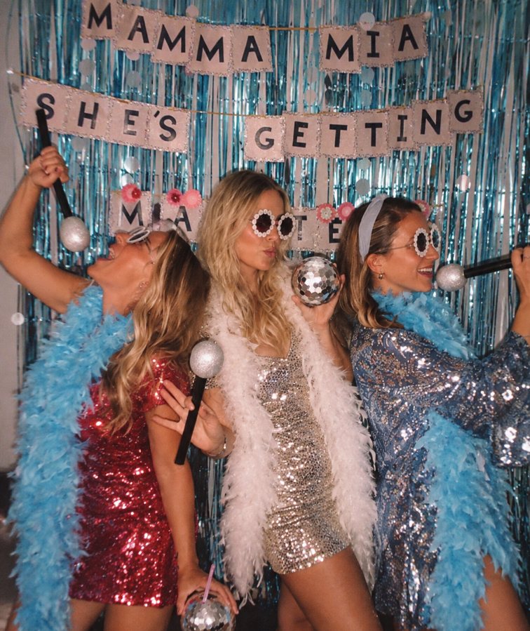 Three friends in sequins and feather boas singing into disco-ball microphones in front of a blue tinsel wall and a Mamma Mia She's Getting Married banner