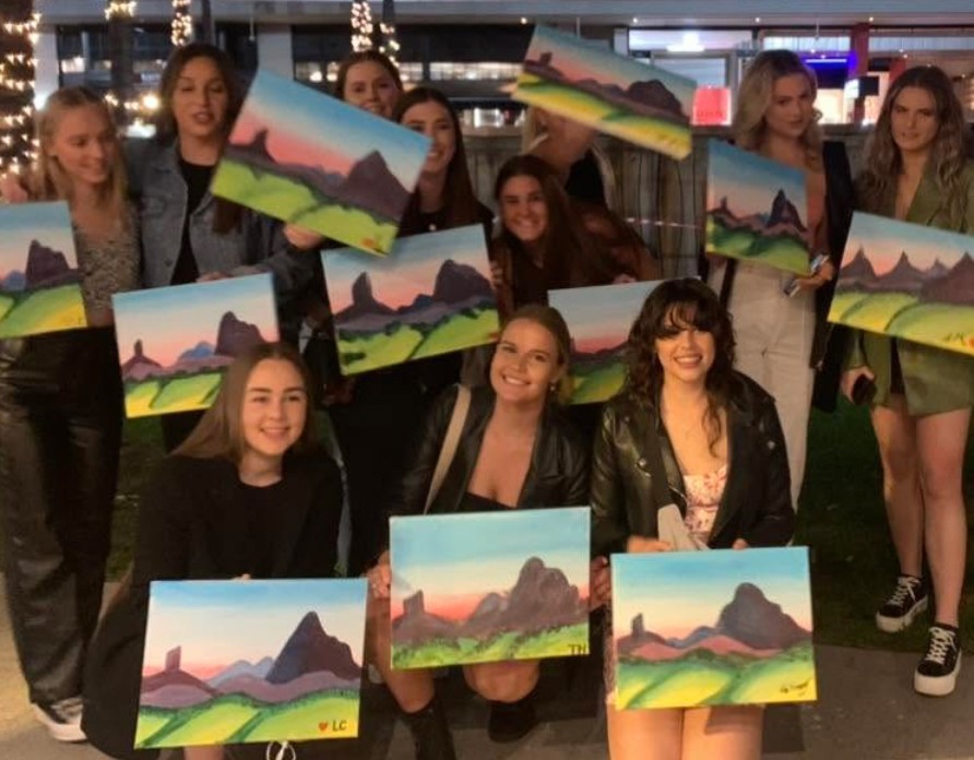 A big crew holding up their mountain paintings outside under fairy lights at night, ready for the rest of the night