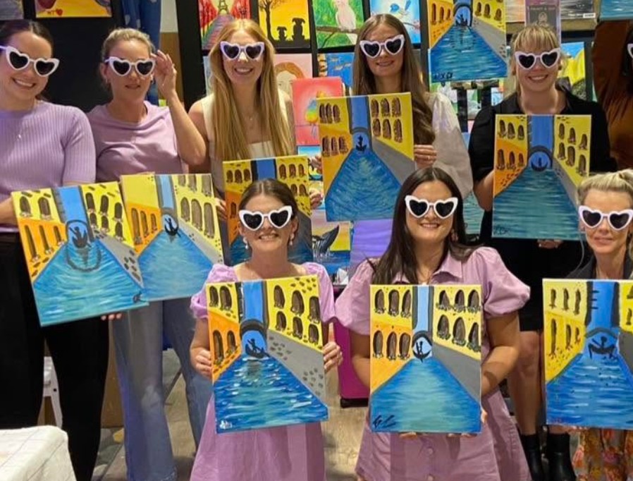A crew in white heart sunglasses holding up their matching canvases of a Venice canal