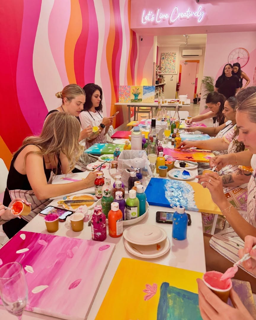 A long table of hens painting bright canvases, a pink and orange mural behind them