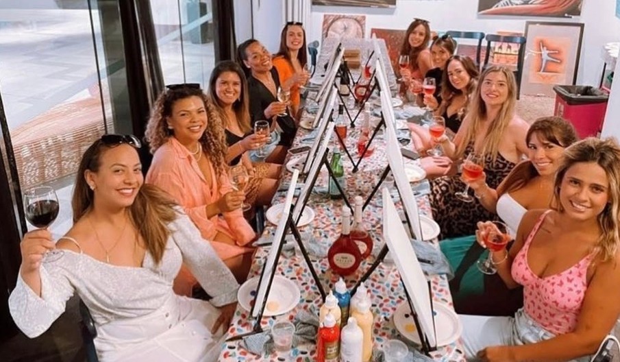 A long table of friends raising glasses of rose and red at their easels before the painting starts
