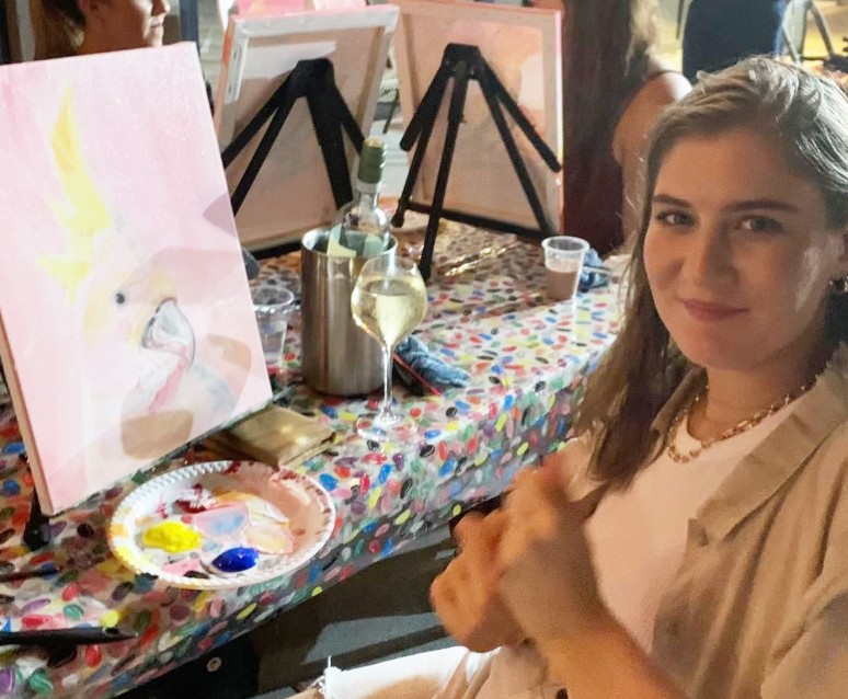 A guest beside her soft pink cockatoo painting, a glass of white in hand