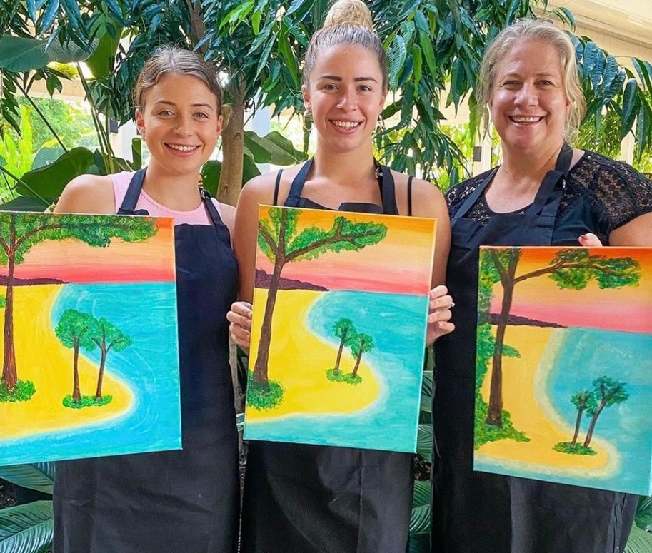Three women of different generations in aprons, smiling and holding up their beach paintings