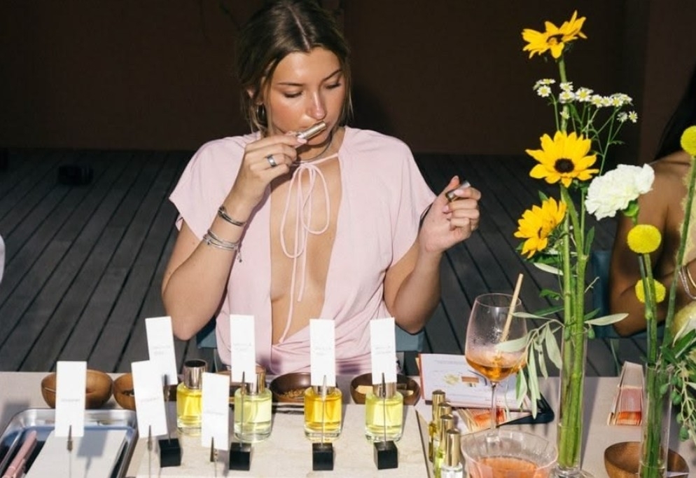 A hen in pink smelling a scent strip at a table of perfume bottles and sunflowers