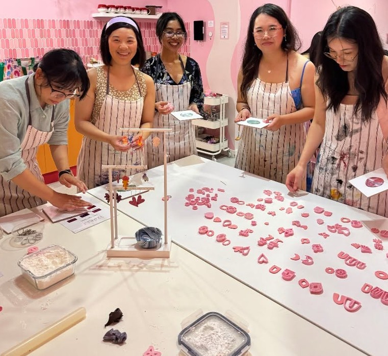 Friends in striped aprons over a table of pink clay earring shapes and an earring stand
