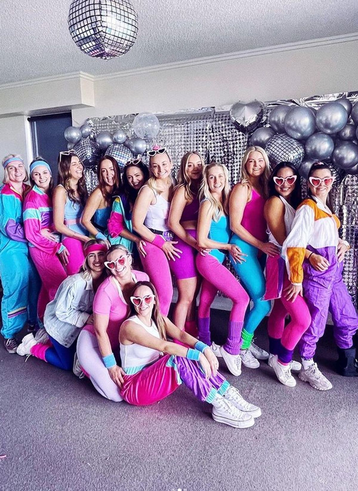A hens group in bright retro outfits posing in front of a silver balloon wall