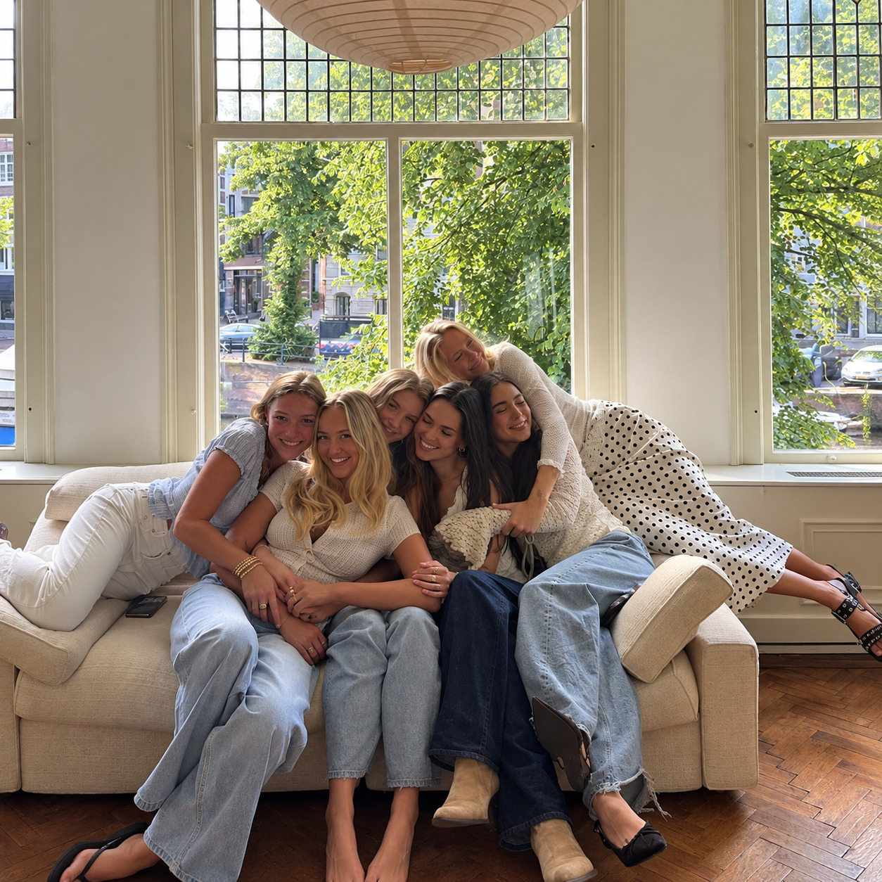 Six friends piled together on a cream couch, laughing and hugging in front of tall windows