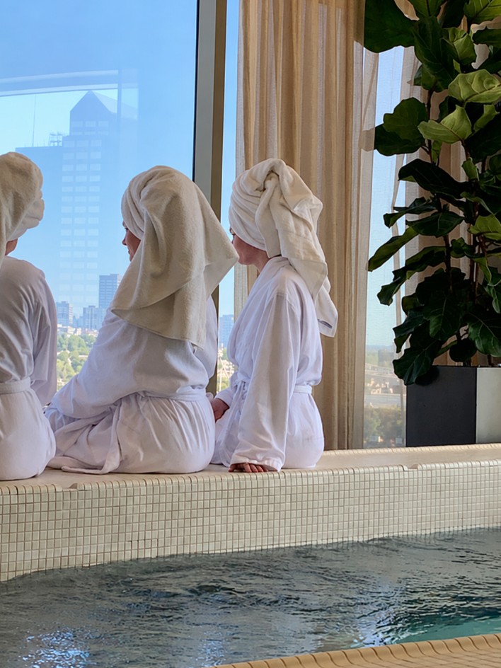 Hens in robes and towel turbans sitting on the edge of a warm pool, looking out a big window over the city