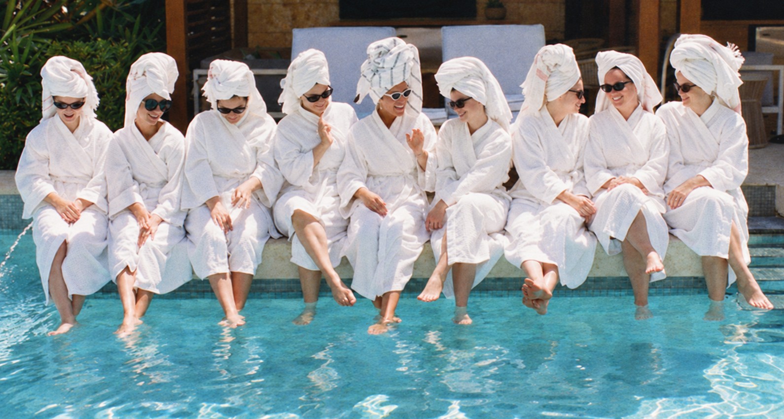 A row of hens in white robes, towel turbans and sunglasses sitting on the edge of a warm pool with their feet in the water
