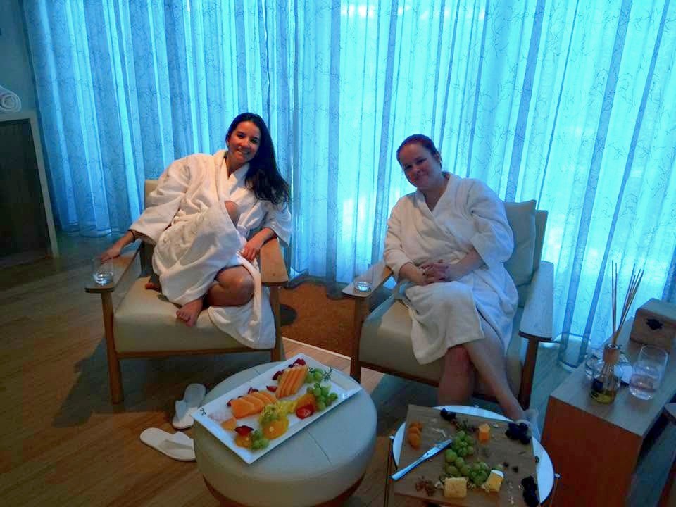 Two friends in robes in armchairs in a blue-curtained lounge, a fruit platter and a cheese board in front of them