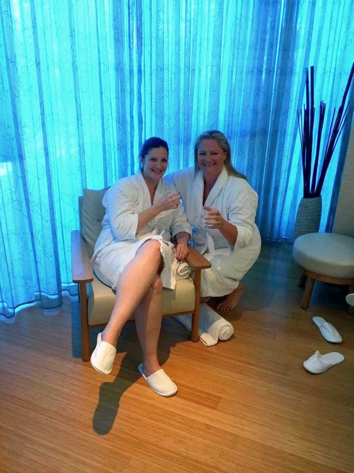 Two friends in robes and slippers sharing an armchair in a quiet relaxation room, glasses raised