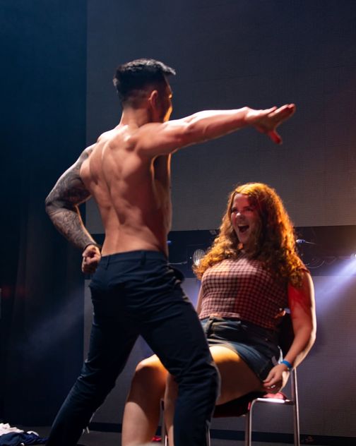 A shirtless performer on stage pointing straight at a hen laughing in the chair