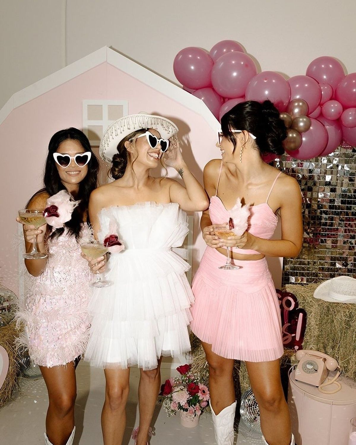 Three friends in pink and white dresses, heart sunglasses and white cowboy boots holding drinks in front of pink balloons