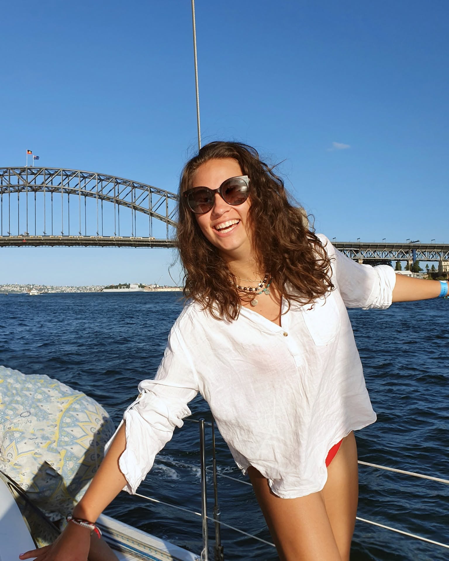 A woman in a white shirt and sunglasses smiling on the bow of a yacht, the harbour bridge behind her