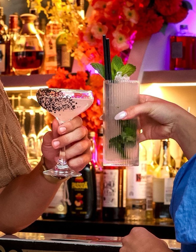 Two friends clinking cocktails at a bar in front of bright flowers