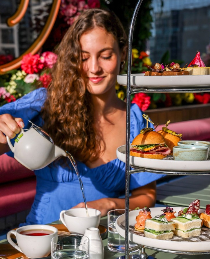 A guest pouring tea beside a three-tier high tea stand of sandwiches, sliders and sweets