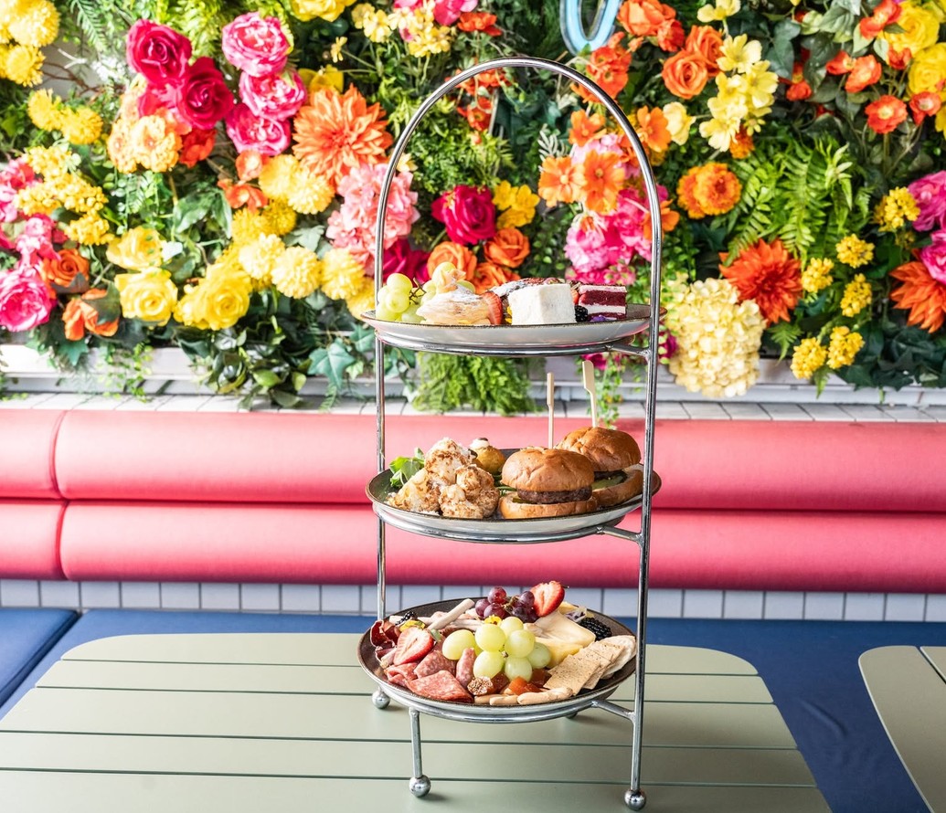 A three-tier high tea stand on a green table in front of a wall of bright flowers