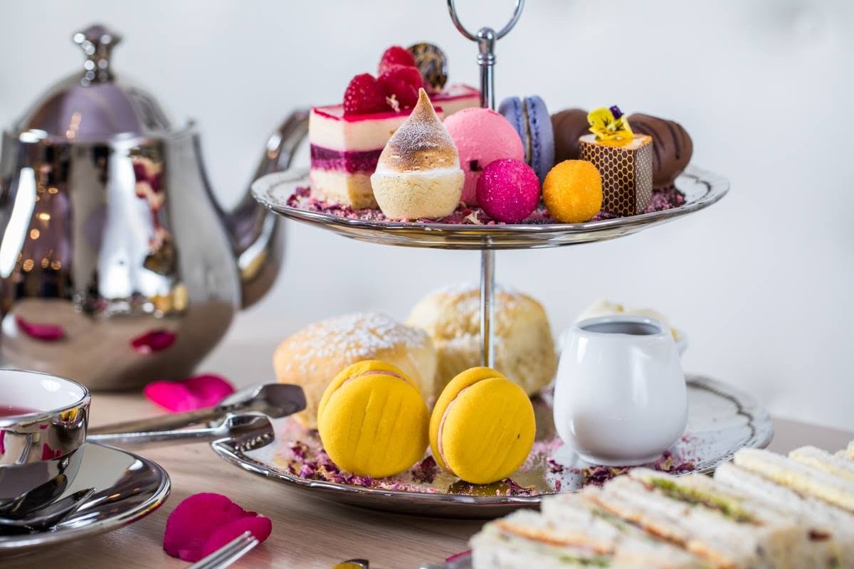 A two-tier stand of little cakes, macarons and scones beside a silver teapot