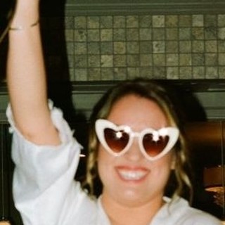 A woman in a white robe and white heart-shaped sunglasses, arm in the air