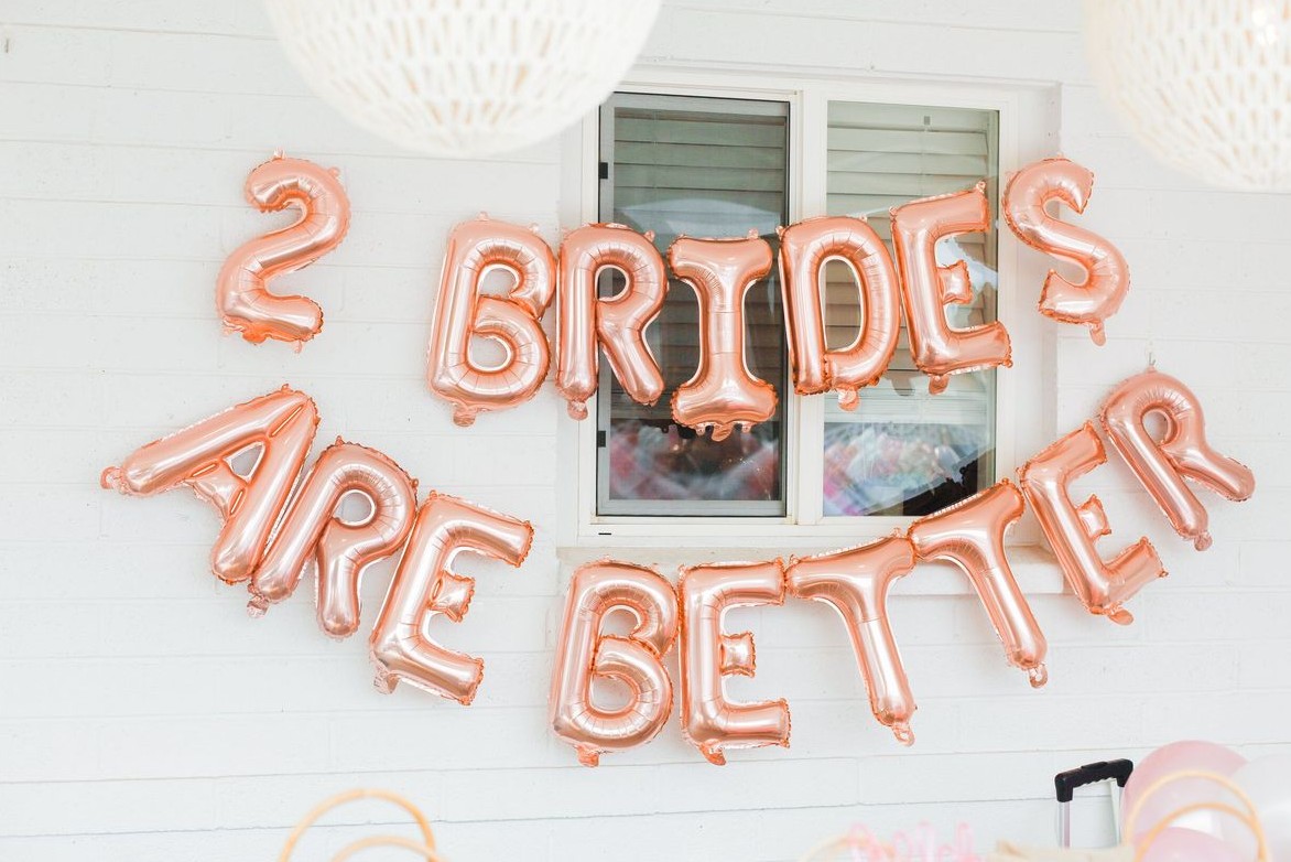 Rose gold balloons spelling 2 Brides Are Better on a white wall