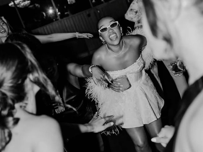 A guest in a white feather dress mid-dance with friends all around, in black and white