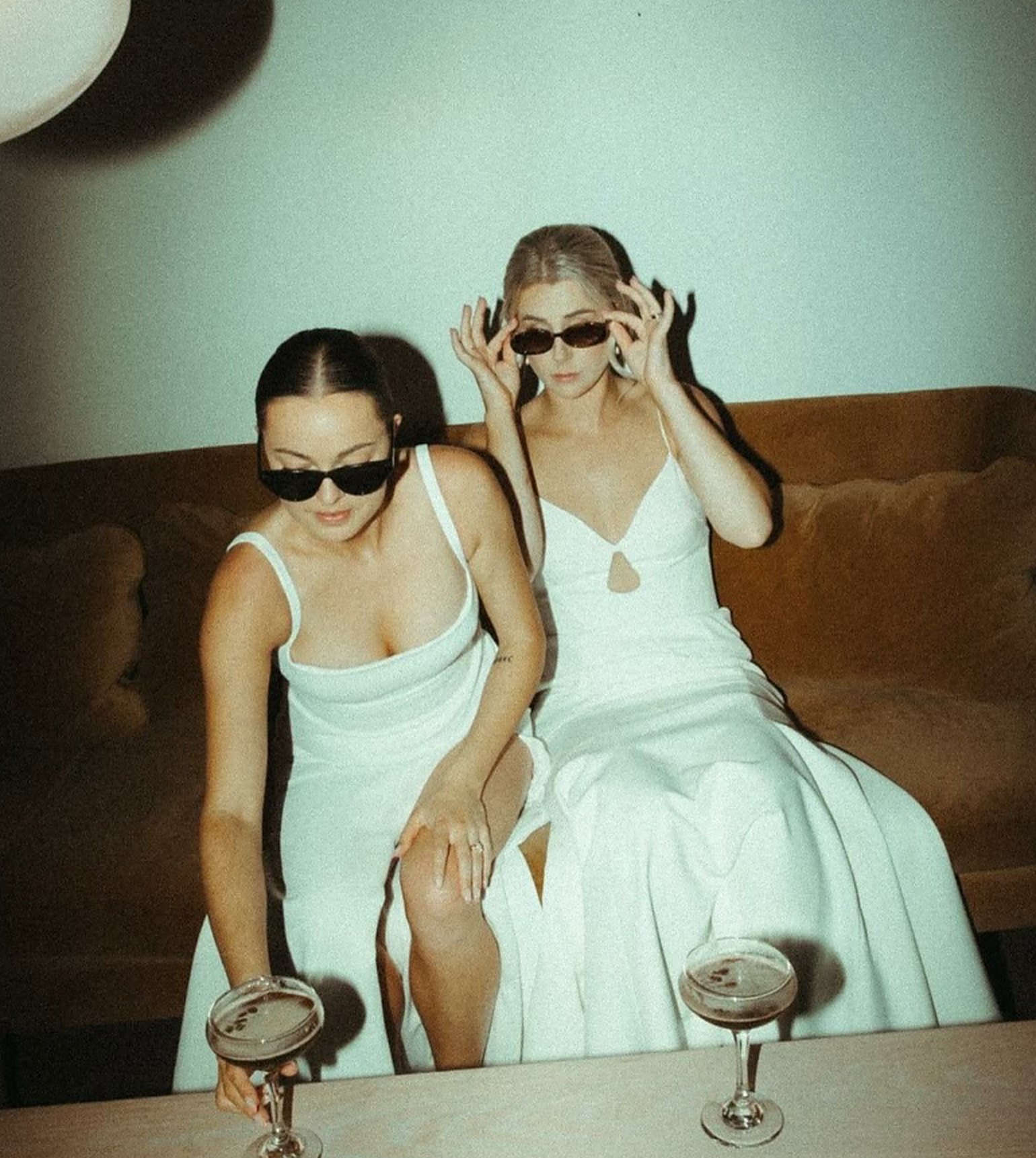 Two people in white dresses lowering their sunglasses on a velvet couch, espresso martinis on the table