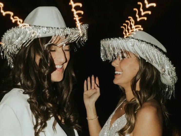 Two guests in sparkling fringed cowboy hats laughing, one in a white suit and one in silver sequins